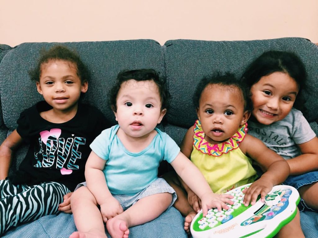 Four toddlers seated on a couch smiling together. The toddlers are children of DLC students.
