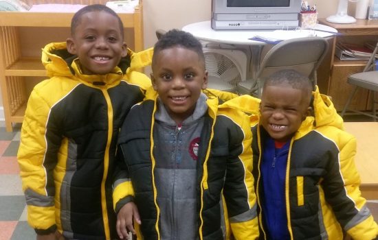 Three young boys in their winter coats smiling together. The young boys are children of DLC students.